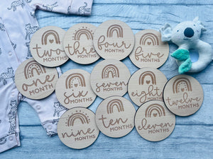 A set of wooden baby milestone discs with rainbow designs, each marked with different ages from one to twelve months, laid out next to a baby's clothing and a plush toy.