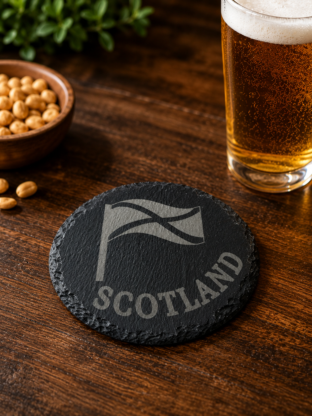 Slate coaster with 'Scotland' and tartan design on a wooden table with a glass of beer and bowl of nuts.