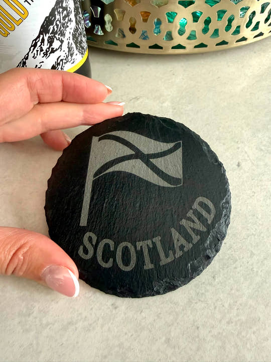 Round black slate coaster with 'Scotland' and a flag design, held by a hand on a light surface.