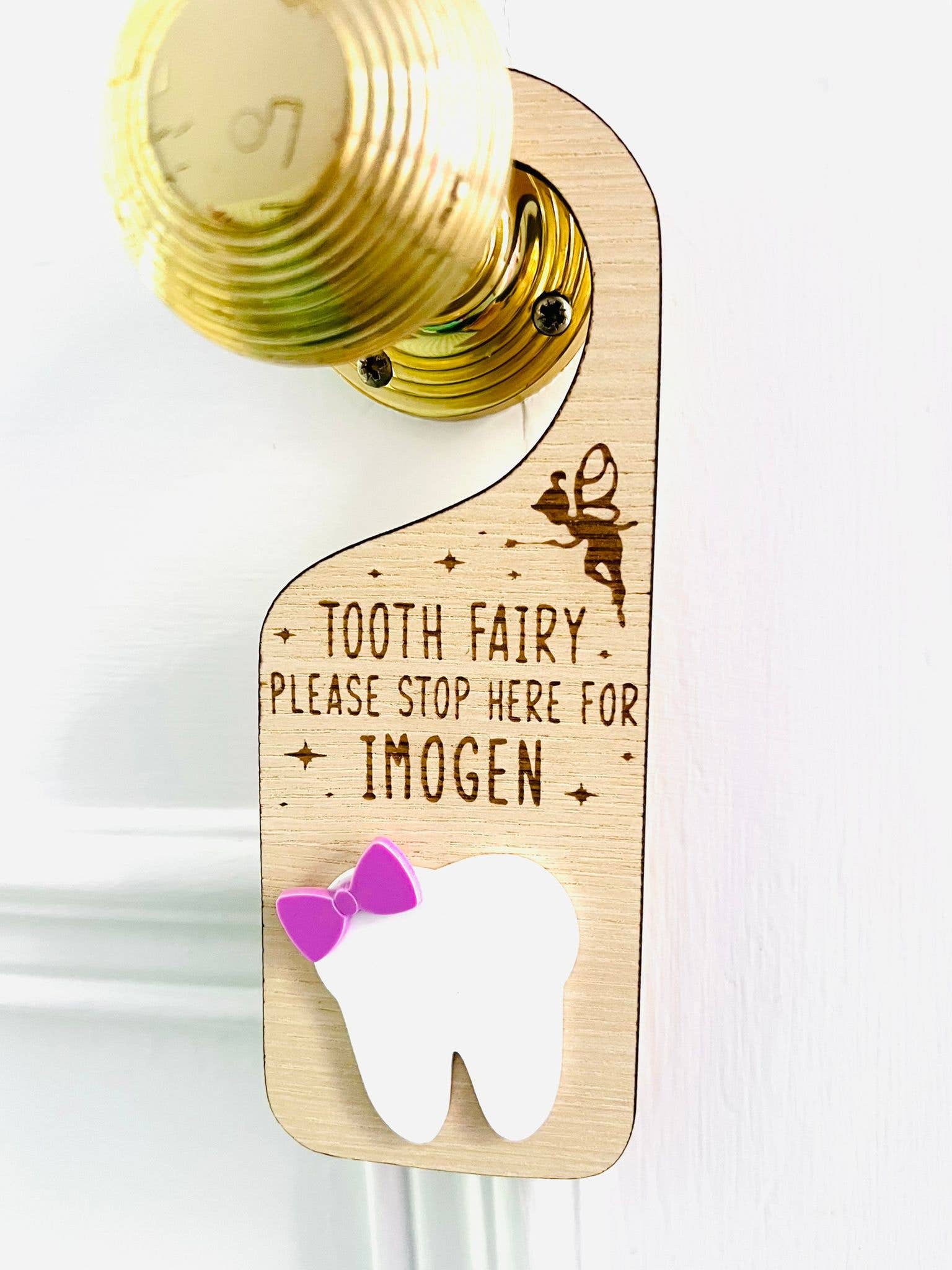 Personalised wooden Tooth Fairy door hanger with engraved text and white tooth shape with purple bow, hanging on a brass doorknob