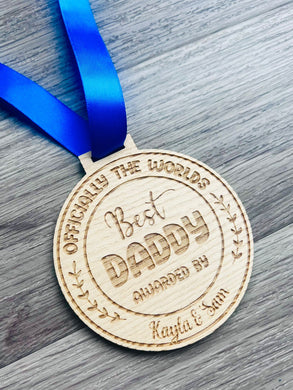 A circular wooden medal engraved with the text “Officially the World’s Best Daddy,” “Awarded By Kayla & Sam,” featuring decorative leaves on the sides. The medal hangs from a royal blue satin ribbon