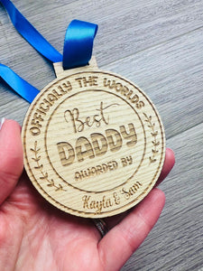 A circular wooden medal engraved with the text “Officially the World’s Best Daddy,” “Awarded By Kayla & Sam,” featuring decorative leaves on the sides. The medal hangs from a royal blue satin ribbon