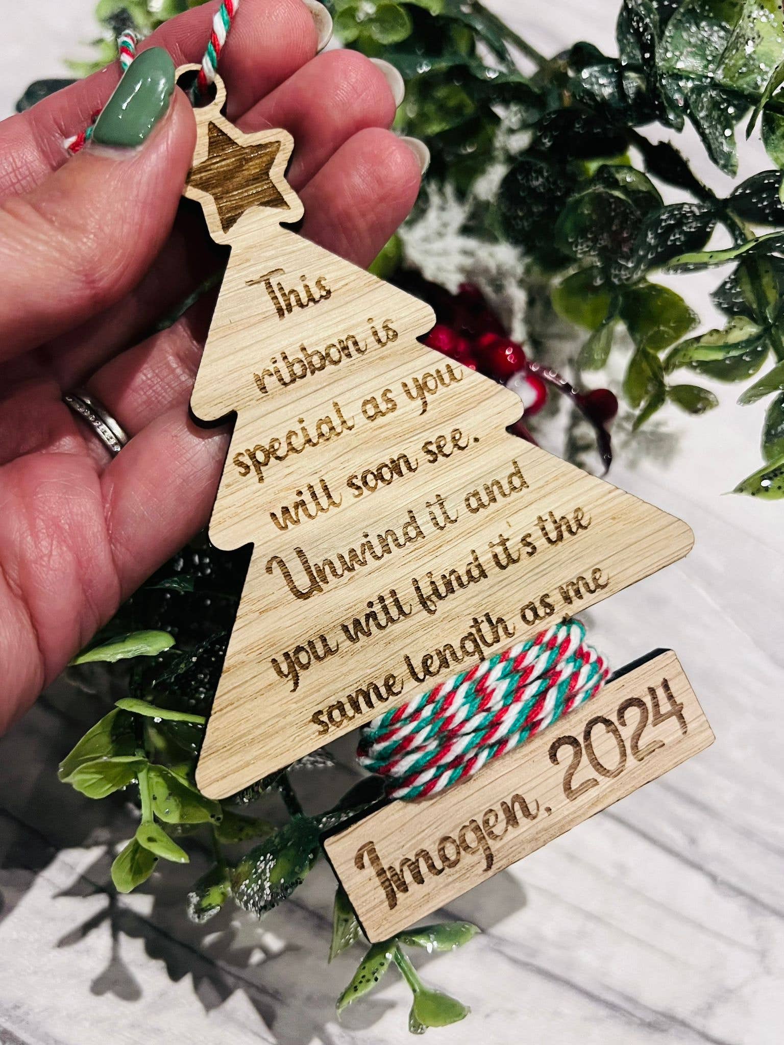 A personalized oak veneer Christmas tree decoration with engraved text, held in a hand, with a background of holly and decorative ribbon.
