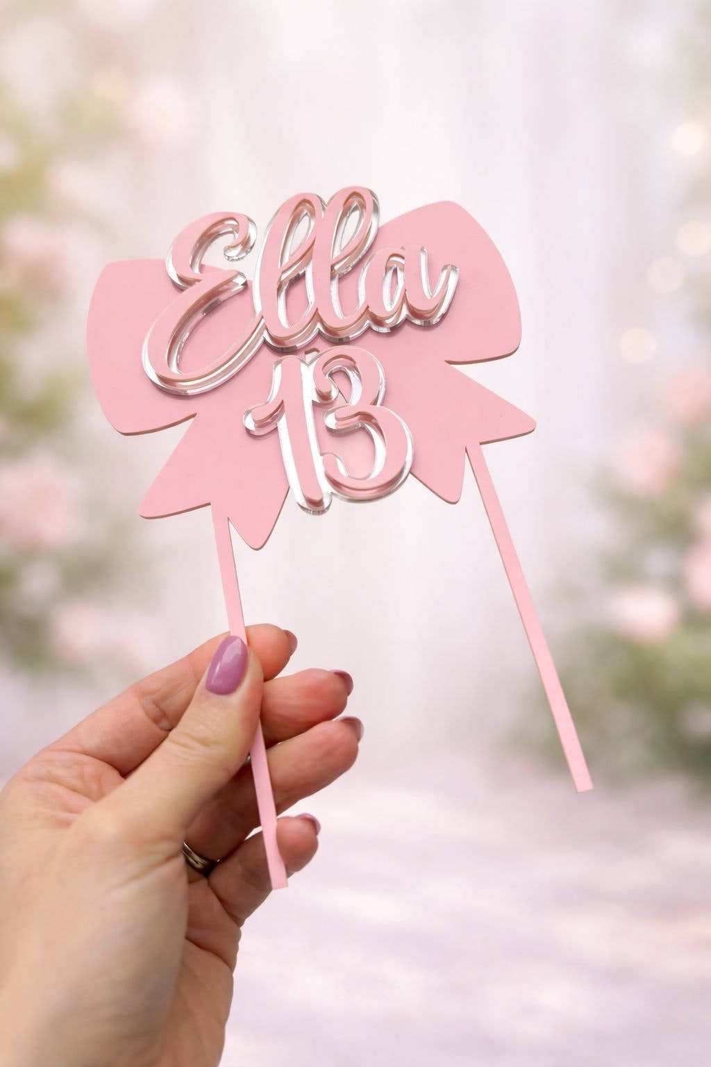 Pink cake topper with 'Ella 13' held by a hand against a blurred background