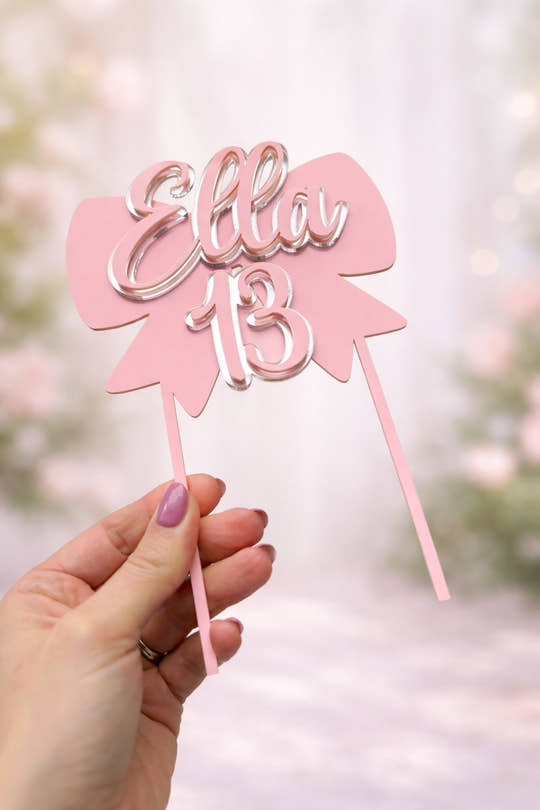 Pink cake topper with 'Ella 13' held by a hand against a blurred background