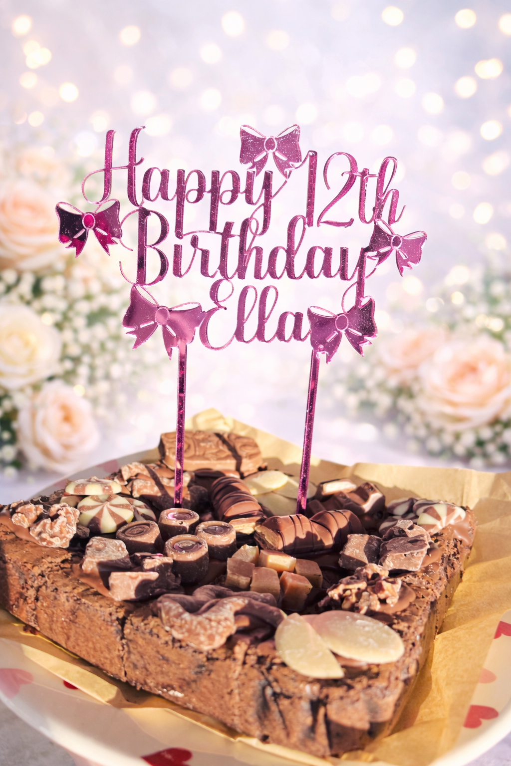 Pink mirrored acrylic personalised cake topper reading “Happy 12th Birthday Ella” displayed on a chocolate brownie celebration cake topped with assorted chocolates against a soft floral background.