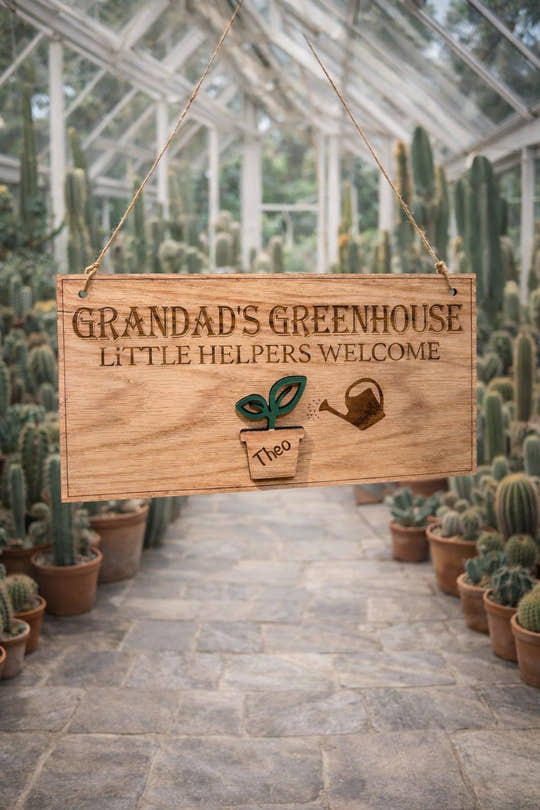 Personalised wooden sign reading “Grandad’s Greenhouse – Little Helpers Welcome” with a plant pot labelled Theo, hanging inside a bright cactus greenhouse with stone flooring.