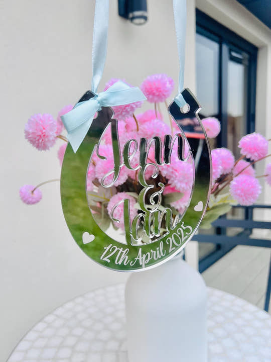 A mirrored horseshoe-shaped wedding decoration with the names "Jenn & Ian" and the date "12th April 2025" engraved. It has a light blue ribbon and bow for hanging, with pink flowers in the background.