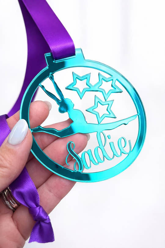 Personalised teal gymnastics medal with name Sadie, held in hand with purple ribbon, against blurred white hall background