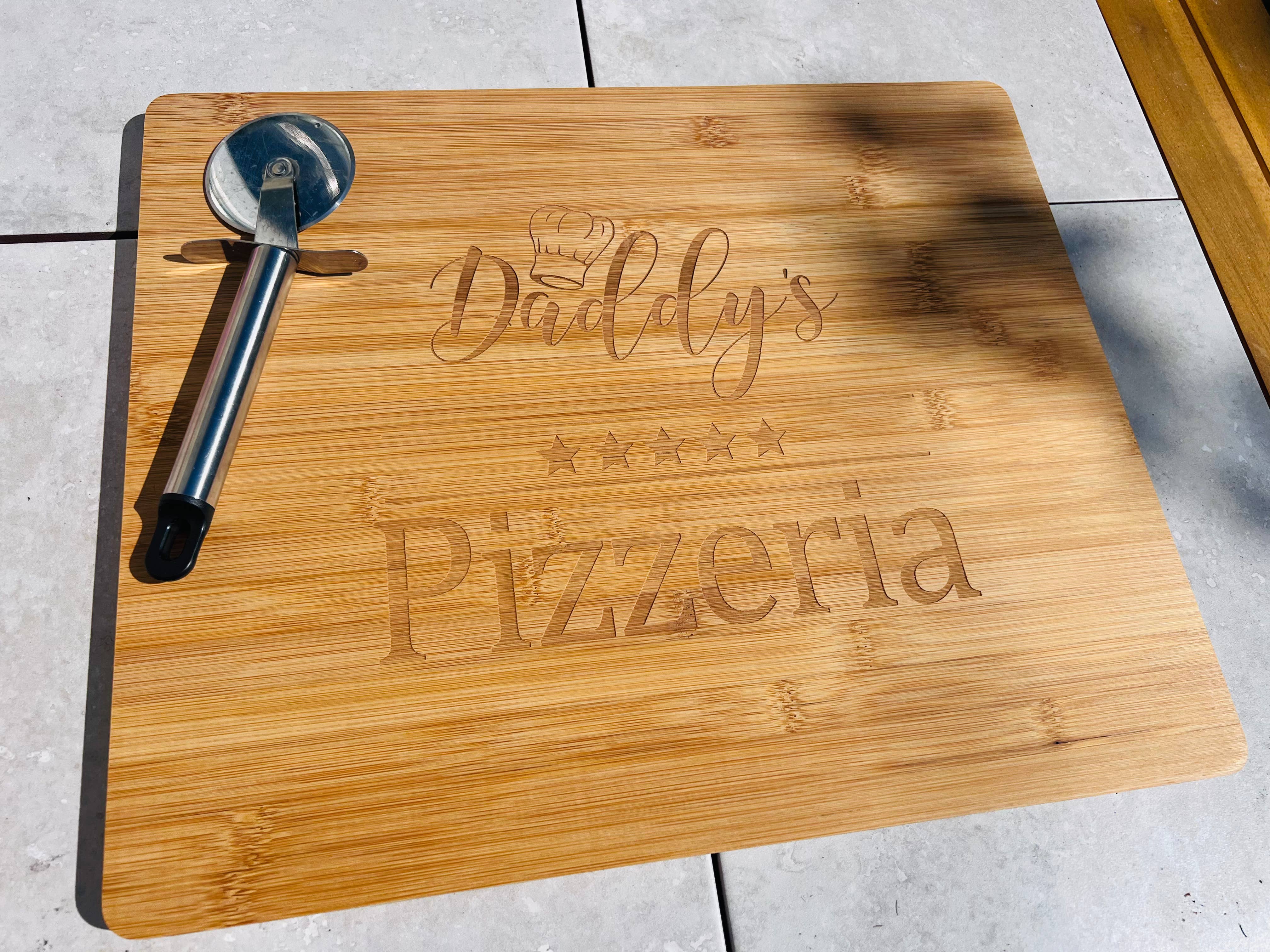 A wooden pizza board with 'Daddy's Pizza' text engraved on it, alongside a metal pizza cutter and a wooden utensil, placed on a tiled surface.