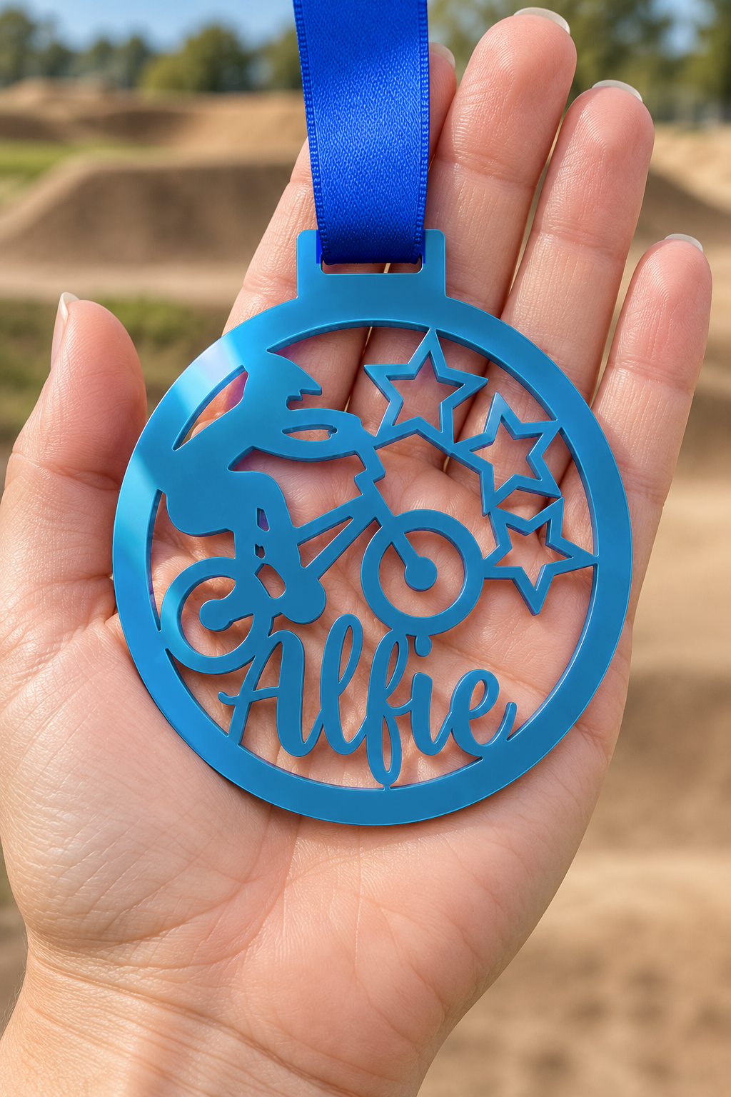 Load image into Gallery viewer, blue mirror acrylic personalised BMX medal with custom name on a blue ribbon, held in hand in front of a blurred BMX track
