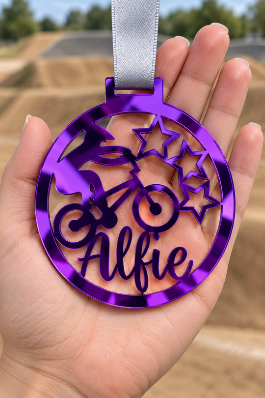 purple mirror acrylic personalised BMX medal with custom name on a silver ribbon, held in hand in front of a blurred BMX track