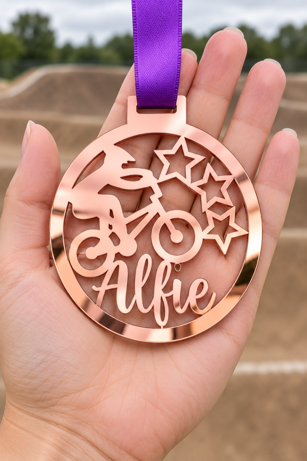 Load image into Gallery viewer, rose gold mirror acrylic personalised BMX medal with custom name on a purple ribbon, held in hand in front of a blurred BMX track
