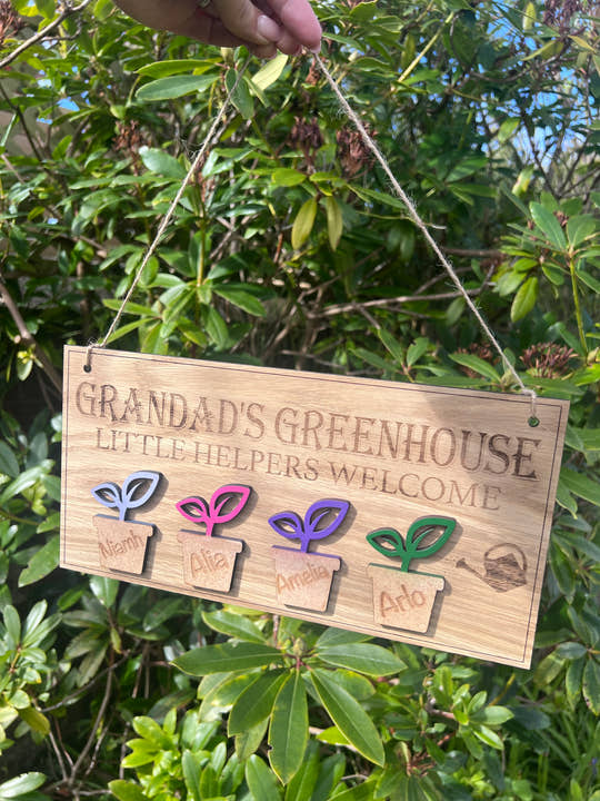 Personalised wooden sign reading “Grandad’s Greenhouse – Little Helpers Welcome” with a plant pot labelled with childrens name, hanging inside a bright garden