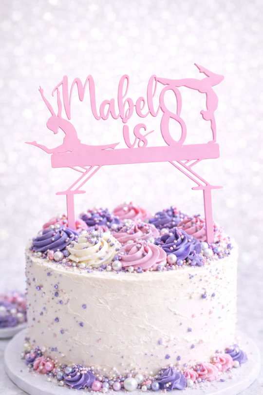 Personalised pink acrylic gymnastics birthday cake topper with custom name and age on white cake decorated with purple and pink buttercream swirls and sprinkles.