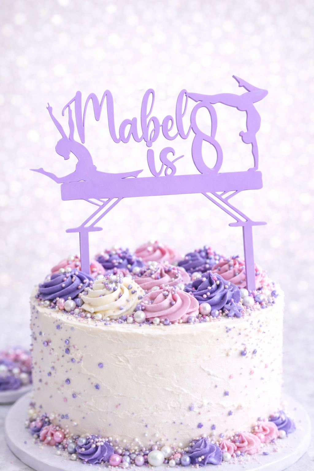 Personalised lilac acrylic gymnastics birthday cake topper with custom name and age on white cake decorated with purple and pink buttercream swirls and sprinkles.