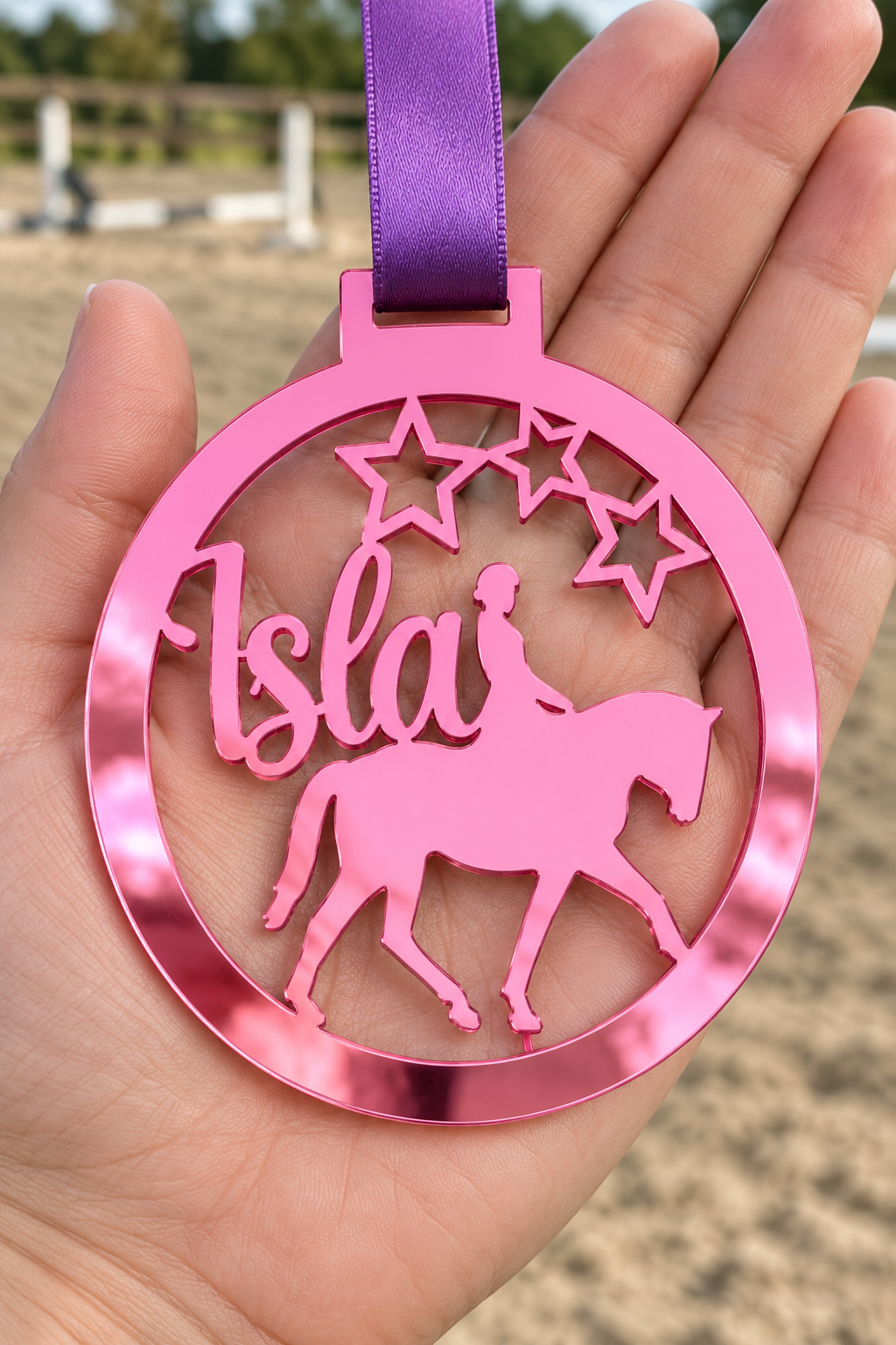 Load image into Gallery viewer, pink mirror acrylic personalised horse riding medal with custom name on a purple ribbon, held in hand in front of a blurred riding arena
