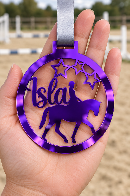 purple mirror acrylic personalised horse riding medal with custom name on a silver ribbon, held in hand in front of a blurred riding arena