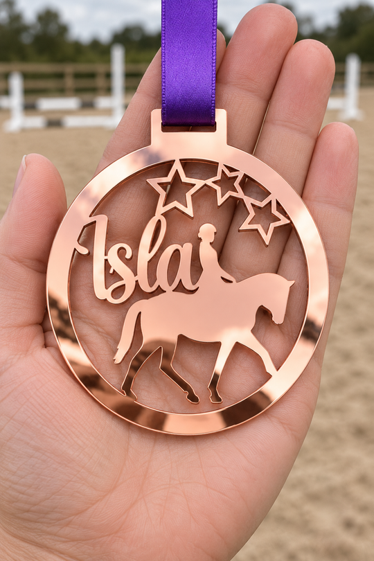 rose gold mirror acrylic personalised horse riding medal with custom name on a purple ribbon, held in hand in front of a blurred riding arena