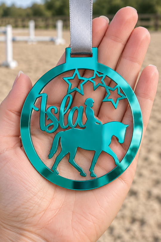 teal mirror acrylic personalised horse riding medal with custom name on a silver ribbon, held in hand in front of a blurred riding arena