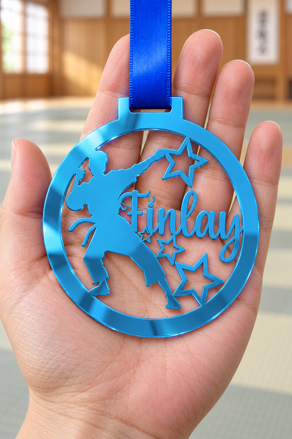 Load image into Gallery viewer, blue mirror acrylic personalised karate boy medal with custom name on a blue ribbon, held in hand in front of a blurred dojo
