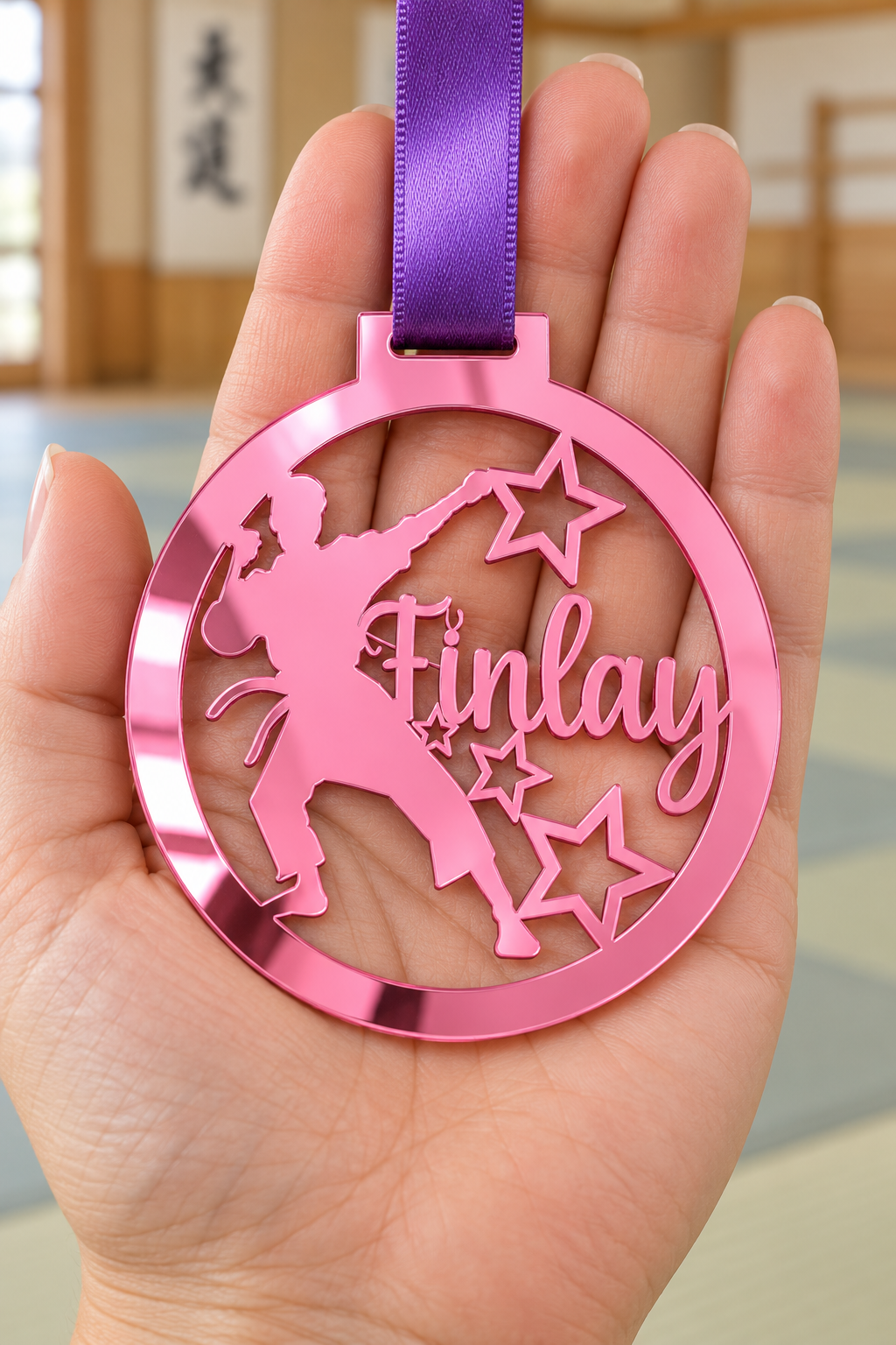 Load image into Gallery viewer, pink mirror acrylic personalised karate boy medal with custom name on a purple ribbon, held in hand in front of a blurred dojo
