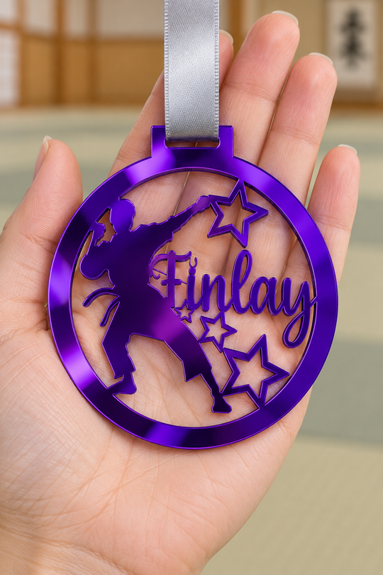 purple mirror acrylic personalised karate boy medal with custom name on a silver ribbon, held in hand in front of a blurred dojo