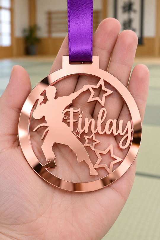 rose gold mirror acrylic personalised karate boy medal with custom name on a purple ribbon, held in hand in front of a blurred dojo