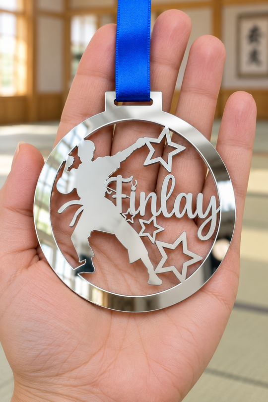 silver mirror acrylic personalised karate boy medal with custom name on a blue ribbon, held in hand in front of a blurred dojo