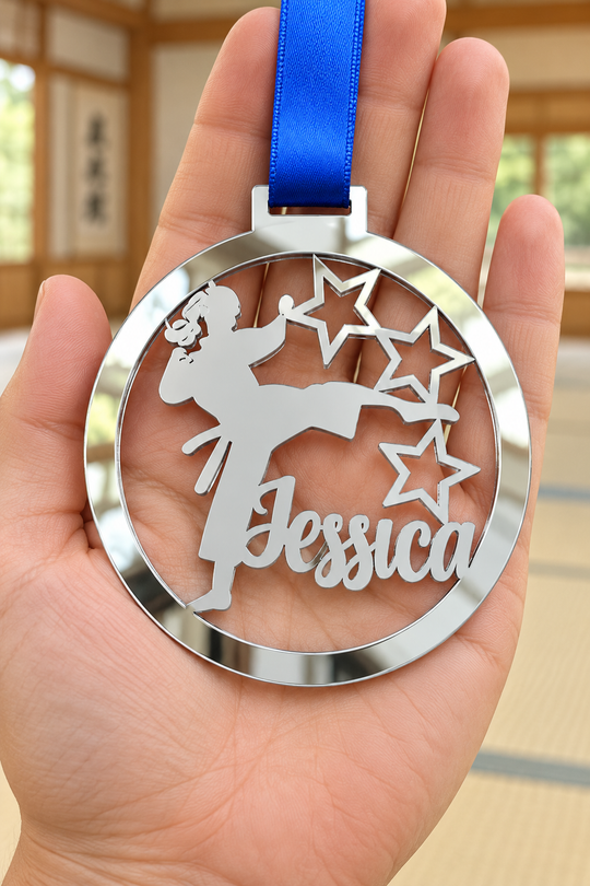 silver mirror acrylic personalised karate girl medal with custom name on a blue ribbon, held in hand in front of a blurred dojo