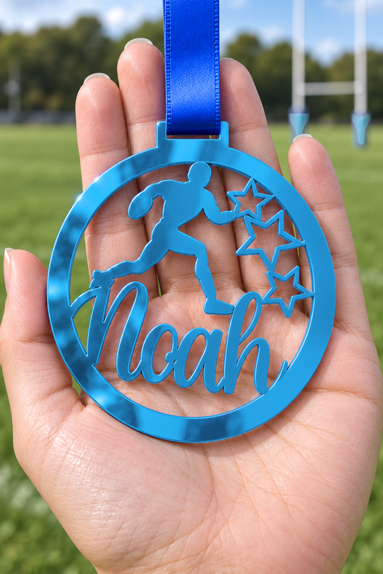 blue mirror acrylic personalised rugby medal with custom name on a blue ribbon, held in hand in front of a blurred rugby pitch