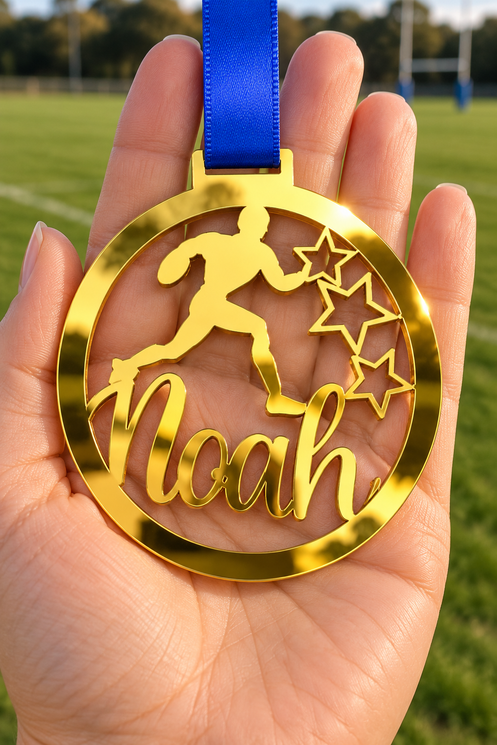 Load image into Gallery viewer, gold mirror acrylic personalised rugby medal with custom name on a blue ribbon, held in hand in front of a blurred rugby pitch
