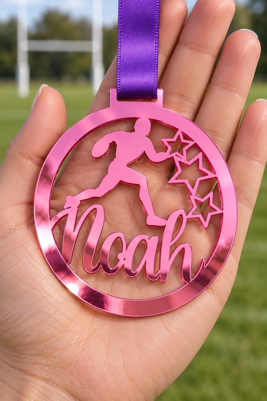 pink mirror acrylic personalised rugby medal with custom name on a purple ribbon, held in hand in front of a blurred rugby pitch