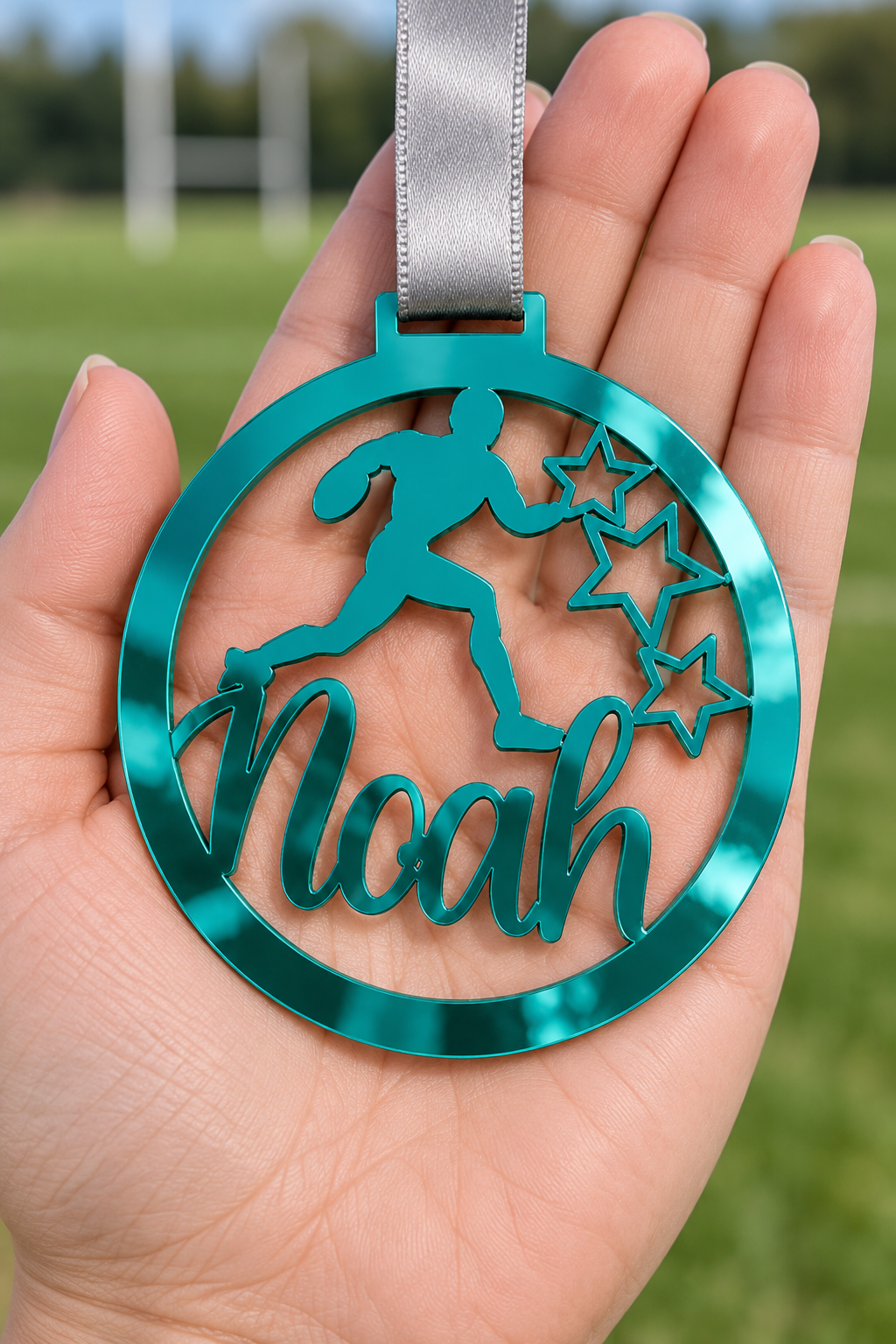 Load image into Gallery viewer, teal mirror acrylic personalised rugby medal with custom name on a silver ribbon, held in hand in front of a blurred rugby pitch
