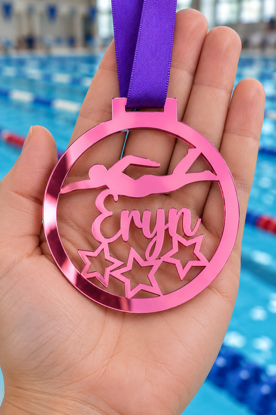 pink mirror acrylic personalised swimming medal with custom name on a purple ribbon, held in hand in front of a blurred swimming pool