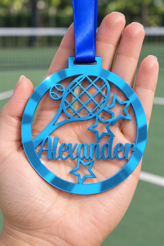 blue mirror acrylic personalised tennis medal with custom name on a blue ribbon, held in hand in front of a blurred tennis court