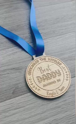A circular wooden medal engraved with the text “Officially the World’s Best Daddy,” “Awarded By Kayla & Sam,” featuring decorative leaves on the sides. The medal hangs from a royal blue satin ribbon
