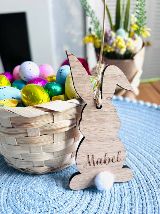 Wooden hanging bunny decoration with the name 'Mabel' inscribed, attached to a Easter tree decoration setup with colorful eggs and Easter-themed items in the background.