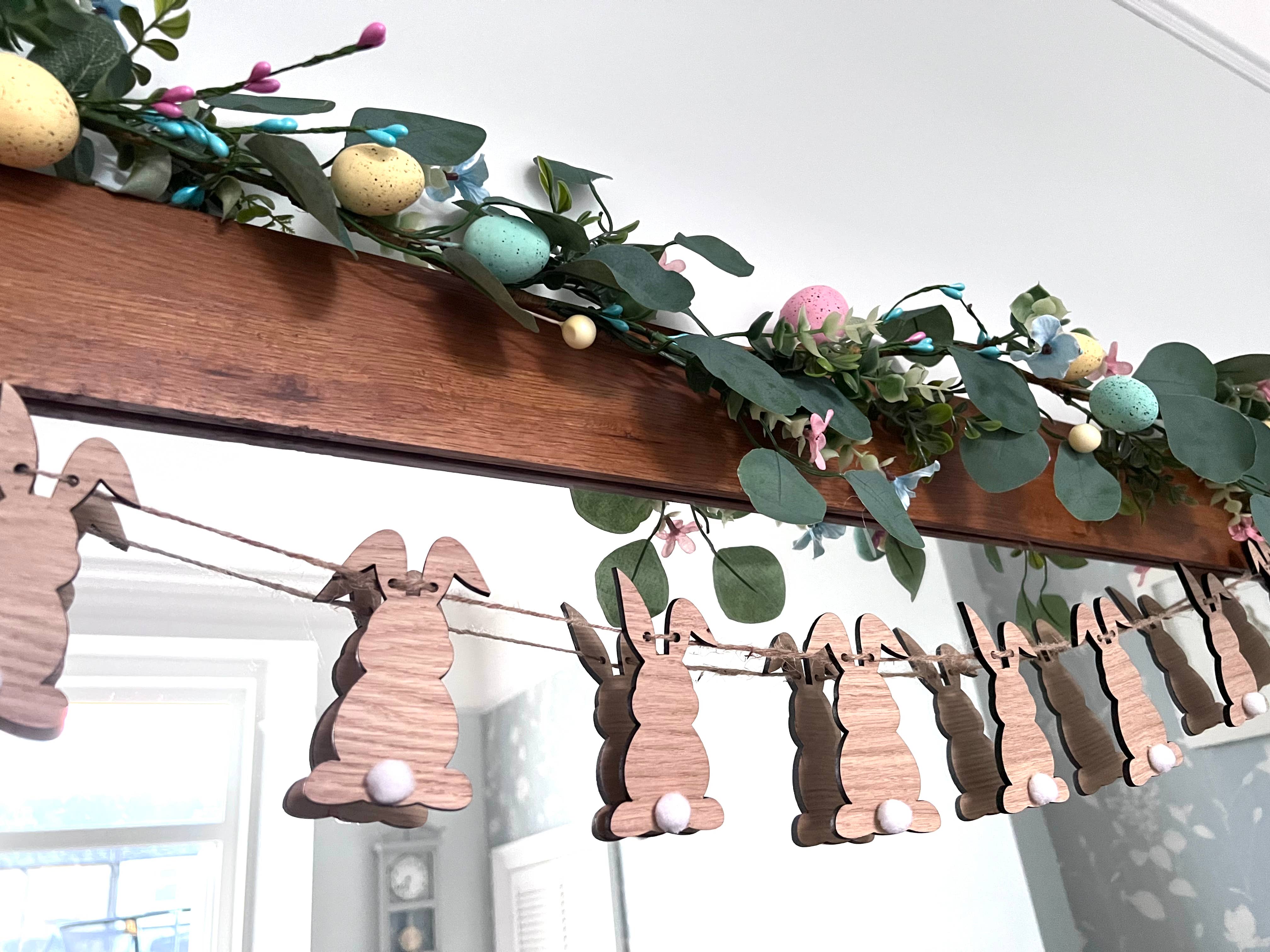 A wooden Easter Bunny Bunting hanging against a wall, featuring laser-cut bunny shapes with white pom-pom tails and decorative foliage.