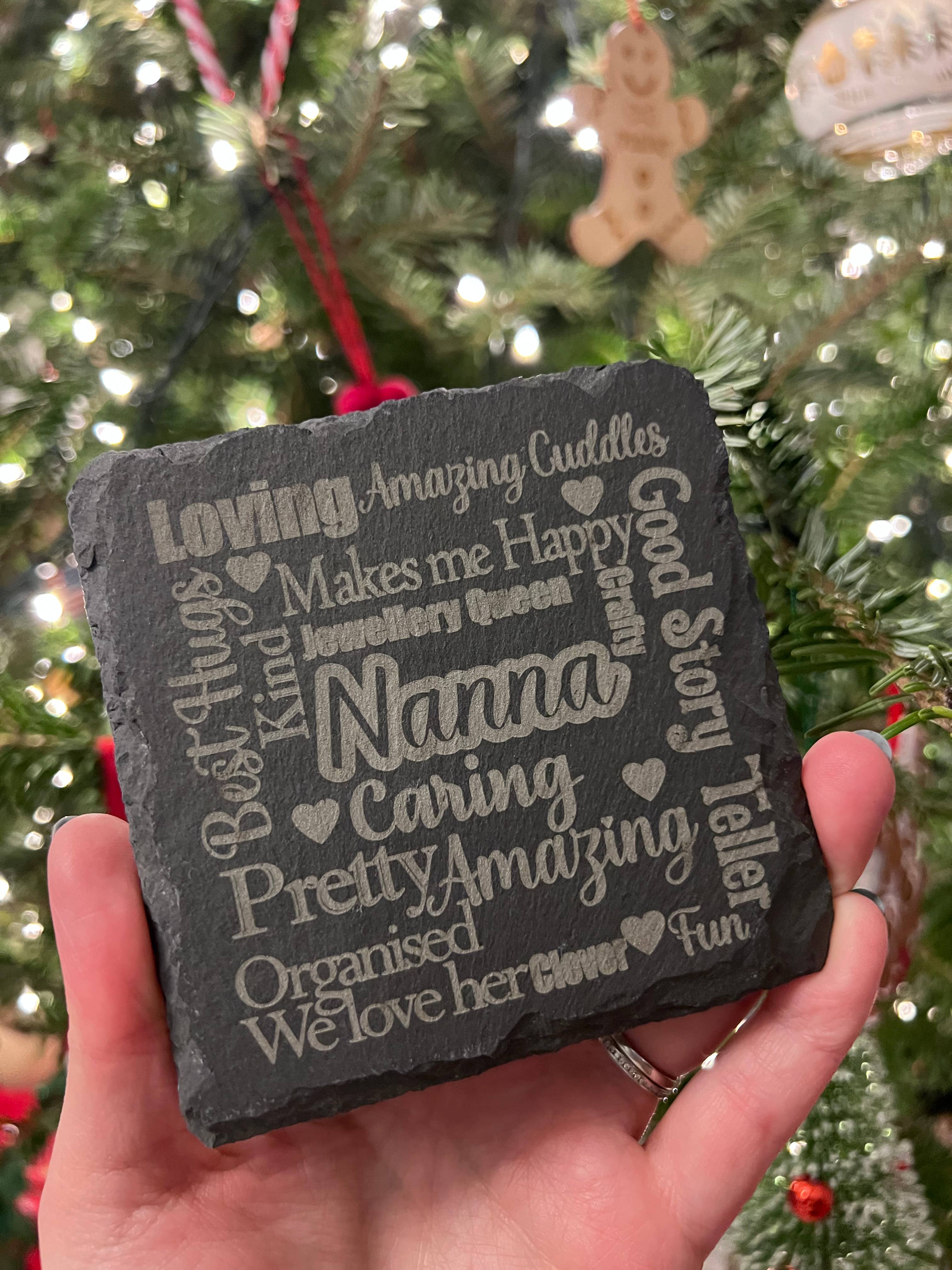 A slate coaster with various words engraved on it, held in a hand with a Christmas tree in the background.
