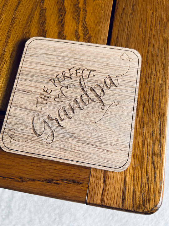 A wooden coaster with 'The Perfect Grandpa' text laser engraved on it, placed on a wooden surface.