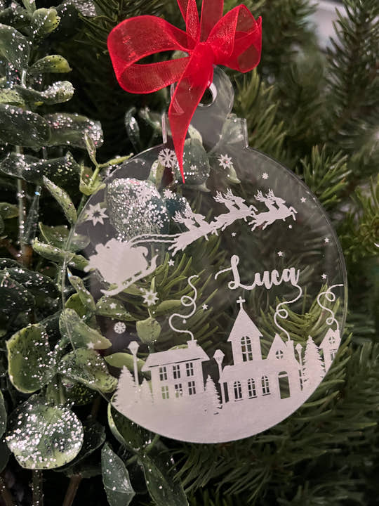 A personalized Christmas decoration featuring a laser-cut and engraved clear acrylic ball with a Christmas scene inside, including a house and snowflakes, with the name 'Lucy' engraved on it, hanging from a red ribbon.