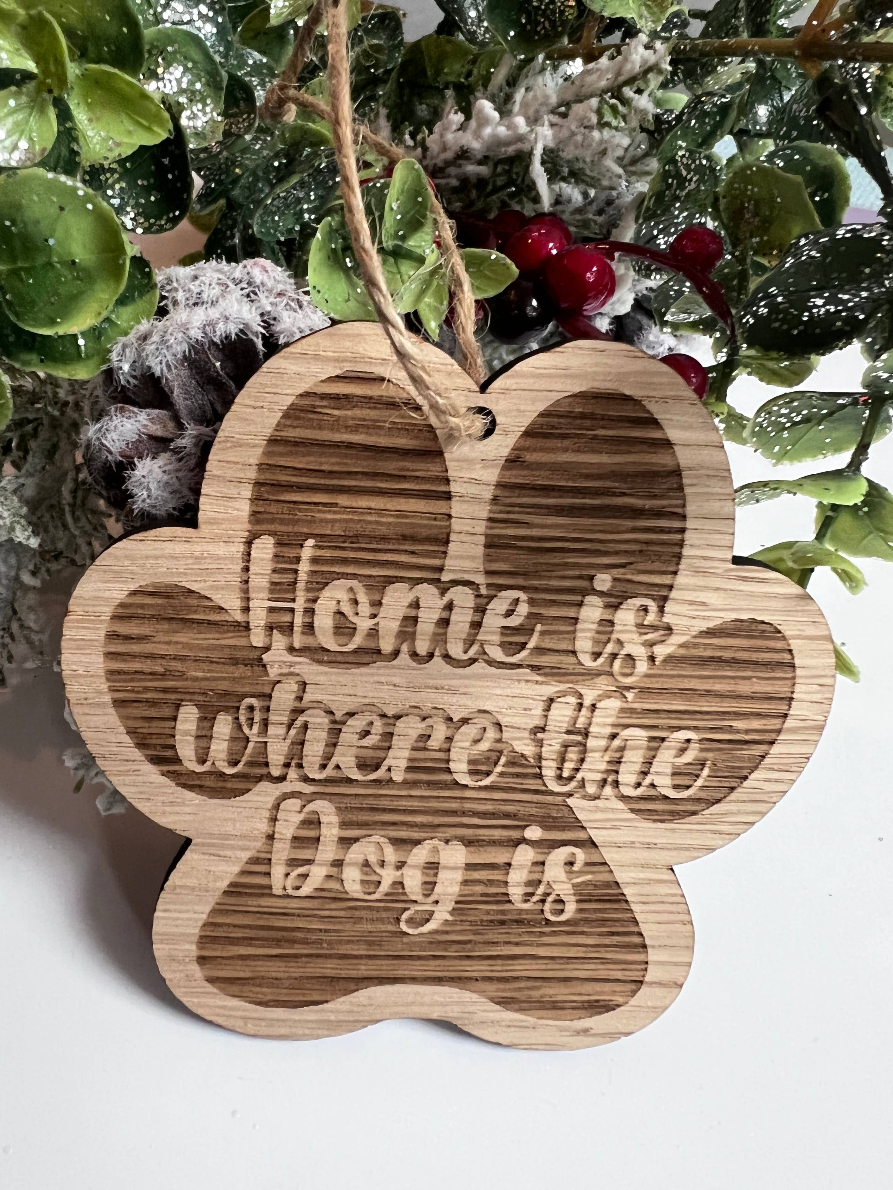 A wooden  paw-shaped decoration with the text 'Home is where the Dog is' engraved on it, displayed against a backdrop of greenery and berries.